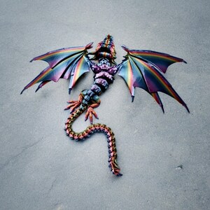 May include: A colourful dragon kite with iridescent wings and a segmented tail. The kite is lying on a grey surface.