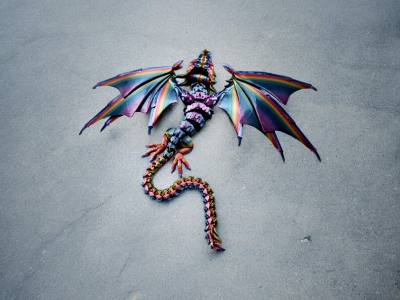 May include: A colourful dragon kite with iridescent wings and a segmented tail. The kite is lying on a grey surface.