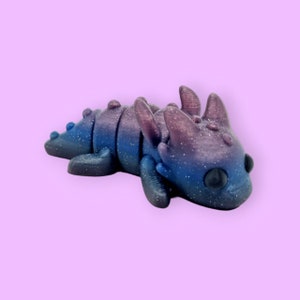 May include: A small, blue and purple dragon figurine with a glittery finish. The dragon is lying on its belly with its head turned to the side. It has large, black eyes and a detailed, textured body.