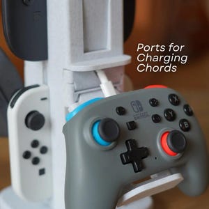 May include: A white and gray charging station for game controllers. The station has multiple slots for controllers and a charging port. A gray Nintendo Switch controller with blue and red accents is in the foreground. Text on the side reads "Ports for Charging Chords."
