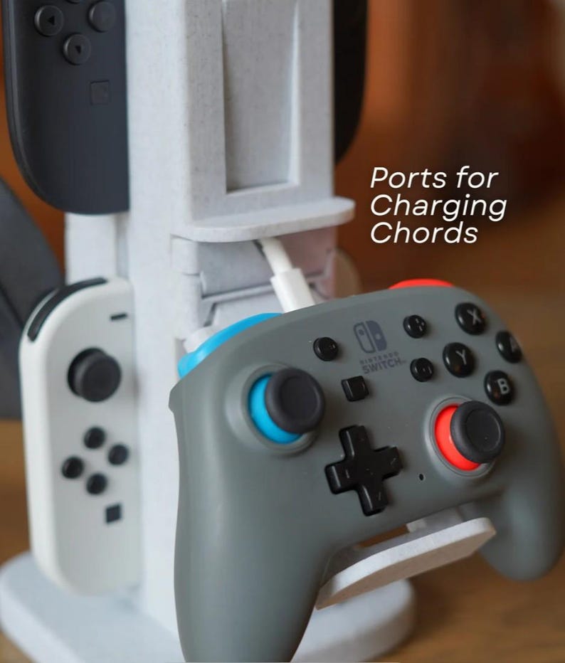May include: A white and gray charging station for game controllers. The station has multiple slots for controllers and a charging port. A gray Nintendo Switch controller with blue and red accents is in the foreground. Text on the side reads "Ports for Charging Chords."
