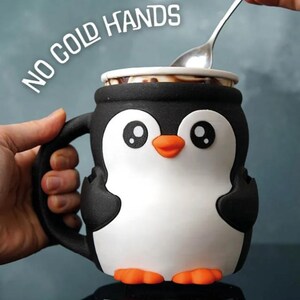 May include: A black, white, and orange penguin-shaped mug with the text "NO COLD HANDS". The mug has a handle and is being filled with a spoon. The penguin has large eyes and orange feet.
