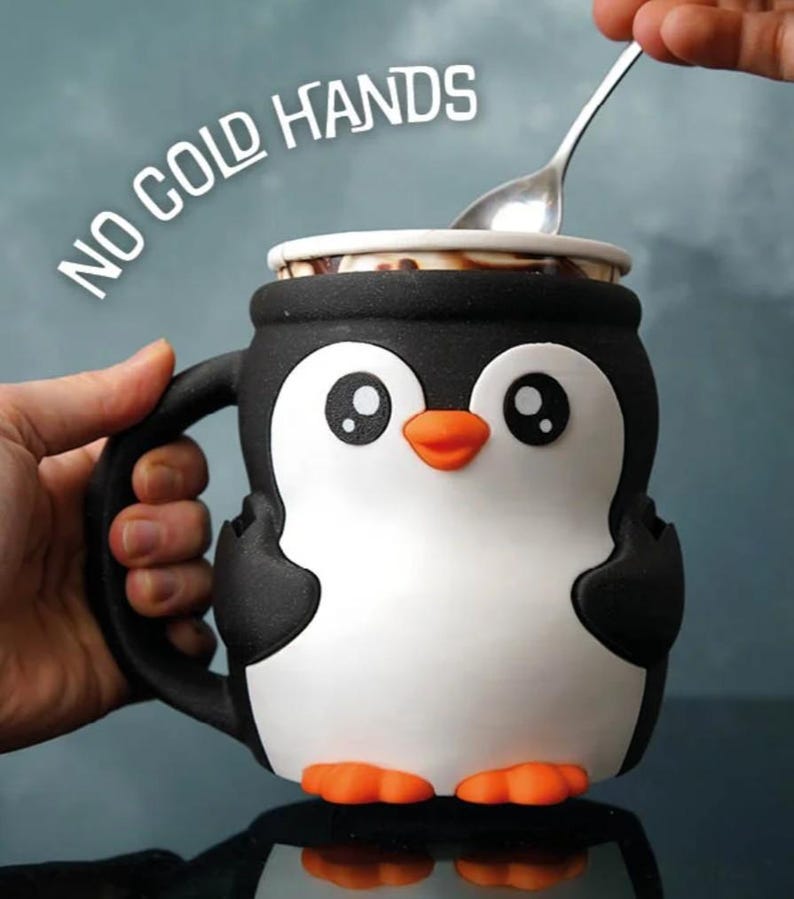 May include: A black, white, and orange penguin-shaped mug with the text "NO COLD HANDS". The mug has a handle and is being filled with a spoon. The penguin has large eyes and orange feet.