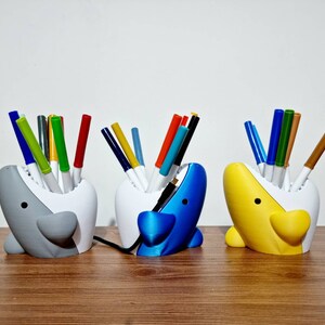 May include: Three shark-shaped pencil holders, each with a different color: gray, blue, and yellow. Each holder has a white mouth and is filled with colorful markers.