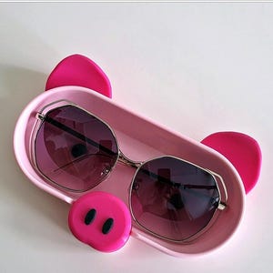 May include: Pink pig-shaped sunglasses case with a pair of black sunglasses inside. The case has a pink snout with two black nostrils.