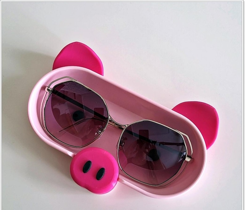 May include: Pink pig-shaped sunglasses case with a pair of black sunglasses inside. The case has a pink snout with two black nostrils.