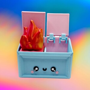 May include: A blue and pink toy box with a red and orange flame on top. The box has a cute cartoon face with two large eyes and a smile.