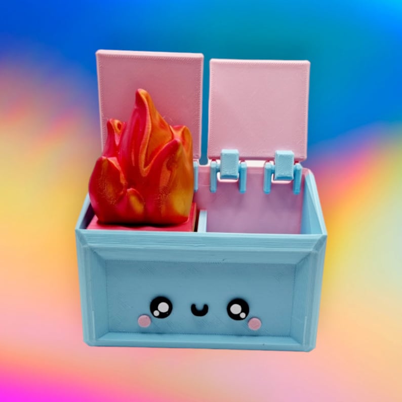 May include: A blue and pink toy box with a red and orange flame on top. The box has a cute cartoon face with two large eyes and a smile.