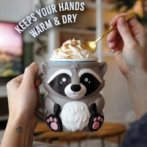 May include: A gray raccoon-shaped mug with black and white details, holding whipped cream topped with cinnamon. The mug has the text "KEEPS YOUR HANDS WARM & DRY" and is held by a person with a gold spoon.