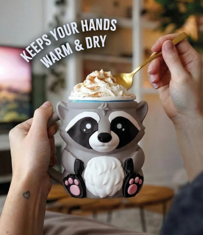 May include: A gray raccoon-shaped mug with black and white details, holding whipped cream topped with cinnamon. The mug has the text "KEEPS YOUR HANDS WARM & DRY" and is held by a person with a gold spoon.