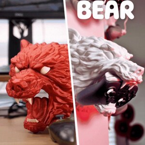 May include: A red and white bear-shaped gaming controller holder. The red bear has an open mouth with visible teeth. The white bear holds a black controller in its mouth. The word "BEAR" is written in white letters.