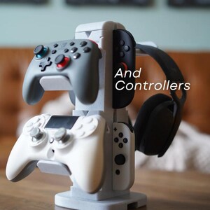 May include: A gray and white gaming controller stand holding two gray controllers, a white controller, and a black headset. The stand has the words "And Controllers" in white text.