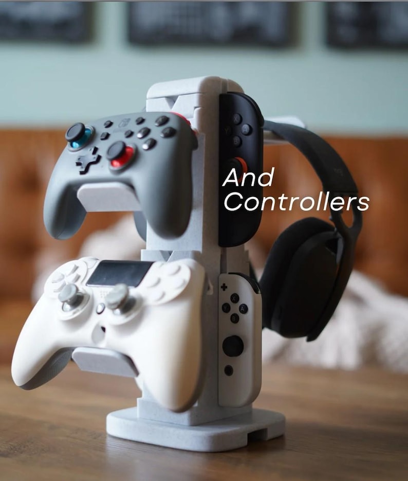May include: A gray and white gaming controller stand holding two gray controllers, a white controller, and a black headset. The stand has the words "And Controllers" in white text.