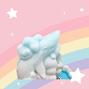 Puede incluir: Una escultura caprichosa, azul claro y blanca, que se asemeja a una nube y un cuerno. La escultura est&aacute; sobre un fondo pastel de arco&iacute;ris y rosa con estrellas blancas. El objeto tiene una textura suave y un dise&ntilde;o l&uacute;dico.