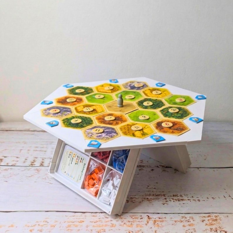 May include: A white, hexagonal board game table with a colorful game board. The game board features various terrain tiles and game pieces. A storage compartment holds game components. The table is supported by angled legs.
