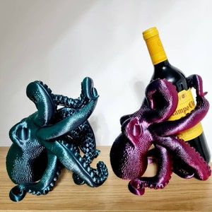 May include: Two 3D printed octopus wine bottle holders. One is teal blue and the other is a deep purple. The purple octopus is holding a bottle of red wine with a yellow label that says "Campo Crianza".