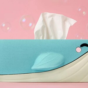 May include: A light blue whale-shaped tissue box with a white base and fin. The whale has a smiling face with a pink cheek and a black eye. White tissues are coming out of the top of the whale. The background is pink with bubbles.