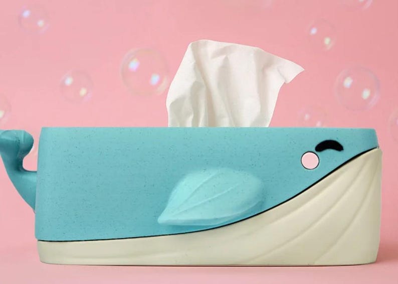 May include: A light blue whale-shaped tissue box with a white base and fin. The whale has a smiling face with a pink cheek and a black eye. White tissues are coming out of the top of the whale. The background is pink with bubbles.