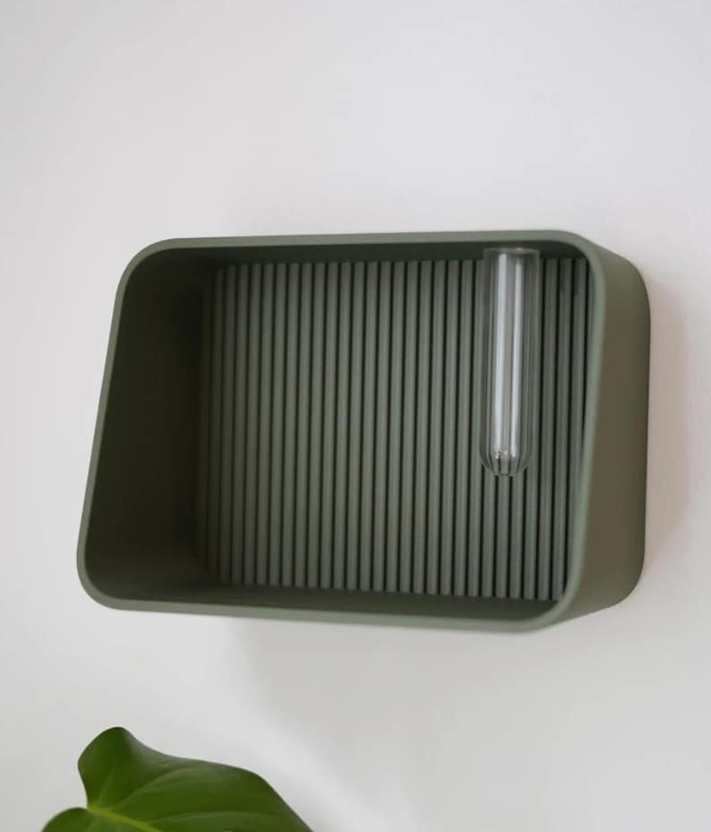 May include: A wall-mounted, olive green planter with a rectangular shape and a ribbed interior. A clear glass test tube is inserted into the planter. The planter is mounted on a white wall. The design is minimalist and modern.