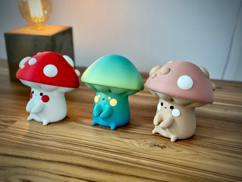 May include: Three small mushroom-shaped figurines with painted details. The mushrooms are red, green, and pink with white spots. The figurines are sitting on a wooden surface.