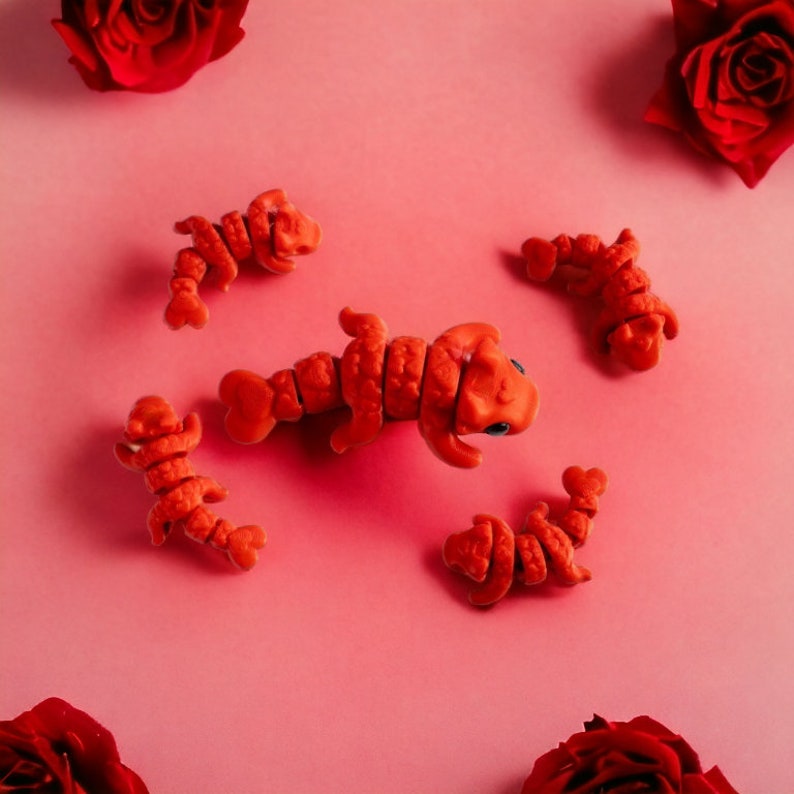 May include: An overhead shot features several orange lobster-shaped candies scattered on a pink surface, with red roses in the corners. The candies have segmented bodies and small blue eyes. The overall theme is playful and whimsical.