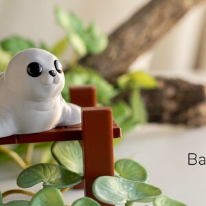 May include: A white baby seal figurine with black eyes sitting on a brown wooden bench. The bench is surrounded by green leafy plants.
