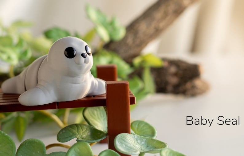 May include: A white baby seal figurine with black eyes sitting on a brown wooden bench. The bench is surrounded by green leafy plants.