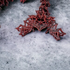 May include: A detailed shot of a red, abstract sculpture with a complex, web-like design. The sculpture is made of a dark red material and rests on a grey, textured surface. The design has an open, intricate structure.