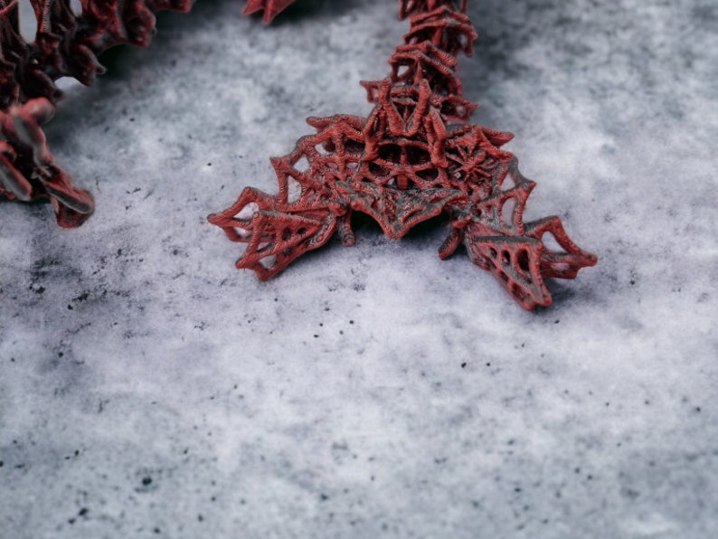 May include: A detailed shot of a red, abstract sculpture with a complex, web-like design. The sculpture is made of a dark red material and rests on a grey, textured surface. The design has an open, intricate structure.