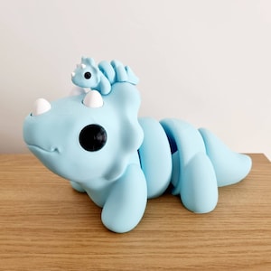 Puede incluir: Un juguete de triceratops articulado, segmentado, de color azul claro con un juguete de triceratops m&aacute;s peque&ntilde;o de color azul claro en la cabeza.