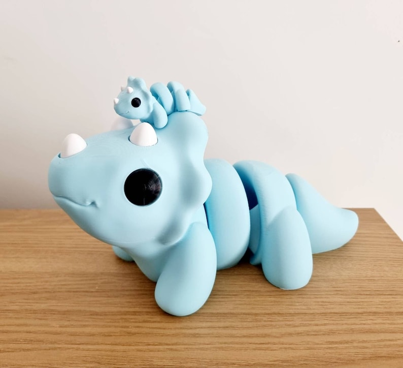 Puede incluir: Un juguete de triceratops articulado, segmentado, de color azul claro con un juguete de triceratops m&aacute;s peque&ntilde;o de color azul claro en la cabeza.