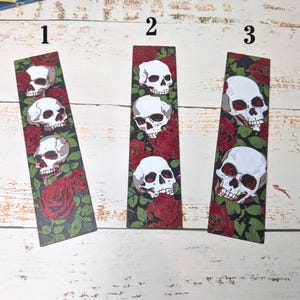 May include: Three rectangular bookmarks with a black background featuring white skulls and red roses. Each bookmark has a different arrangement of skulls and roses. The bookmarks are on a white wooden surface.
