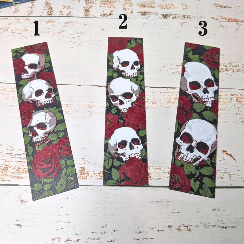 May include: Three rectangular bookmarks with a black background featuring white skulls and red roses. Each bookmark has a different arrangement of skulls and roses. The bookmarks are on a white wooden surface.