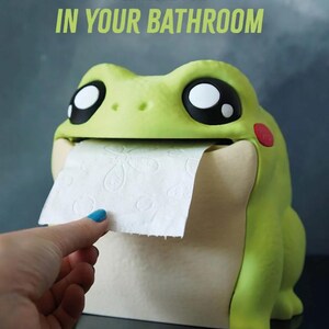 May include: A whimsical frog-shaped toilet paper holder in shades of green and cream. The frog has large, expressive eyes and a red cheek. The text "FOR MORE FUN IN YOUR BATHROOM" is displayed above the frog. A hand is pulling a sheet of toilet paper.