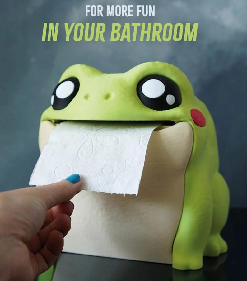 May include: A whimsical frog-shaped toilet paper holder in shades of green and cream. The frog has large, expressive eyes and a red cheek. The text "FOR MORE FUN IN YOUR BATHROOM" is displayed above the frog. A hand is pulling a sheet of toilet paper.