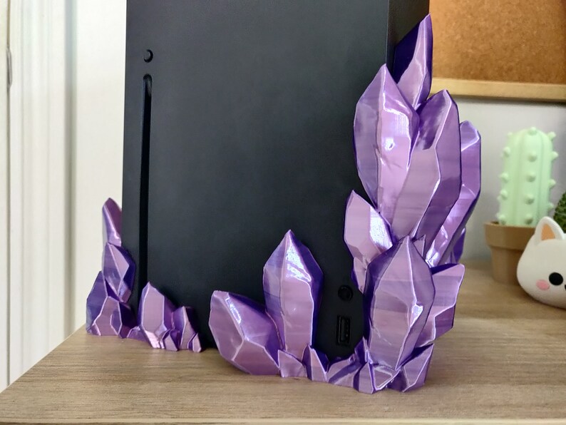 Puede incluir: Una consola de juegos negra con un soporte de cristal morado. El soporte est&aacute; hecho de pl&aacute;stico impreso en 3D y tiene un acabado brillante e iridiscente.