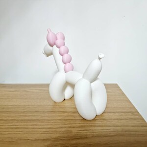 May include: White balloon animal sculpture of a unicorn with a pink mane. The unicorn is standing on a wooden surface.