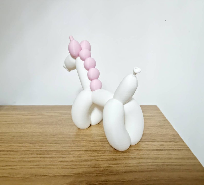 May include: White balloon animal sculpture of a unicorn with a pink mane. The unicorn is standing on a wooden surface.