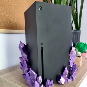 Puede incluir: Una consola de juegos negra con un soporte en forma de cristal morado. La consola est&aacute; sobre una superficie de madera.