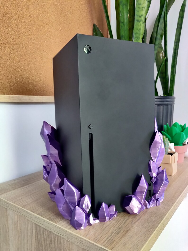 Puede incluir: Una consola de juegos negra con un soporte en forma de cristal morado. La consola est&aacute; sobre una superficie de madera.