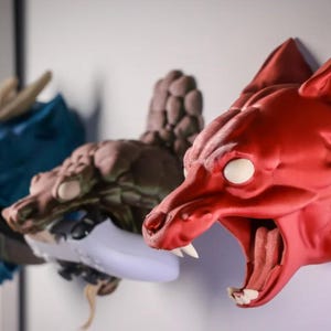 Puede incluir: Tres soportes de pared de cabeza de animal impresos en 3D. El primero es un drag&oacute;n azul, el segundo es un drag&oacute;n marr&oacute;n y el tercero es un lobo rojo con la boca abierta.