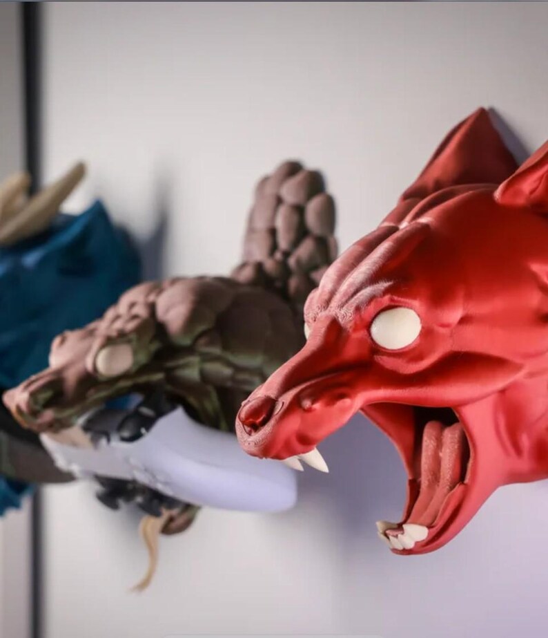 Puede incluir: Tres soportes de pared de cabeza de animal impresos en 3D. El primero es un drag&oacute;n azul, el segundo es un drag&oacute;n marr&oacute;n y el tercero es un lobo rojo con la boca abierta.