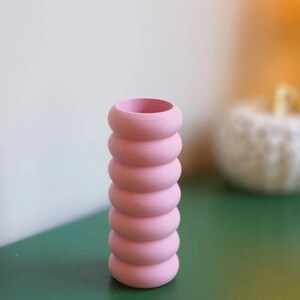May include: A pink vase with a unique, stacked ring design. The vase is a soft, pastel pink and has a cylindrical shape. The vase is approximately 8 inches tall and is sitting on a green surface.