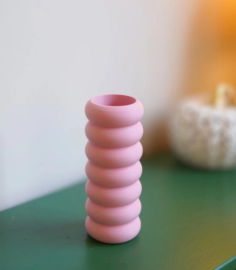 May include: A pink vase with a unique, stacked ring design. The vase is a soft, pastel pink and has a cylindrical shape. The vase is approximately 8 inches tall and is sitting on a green surface.
