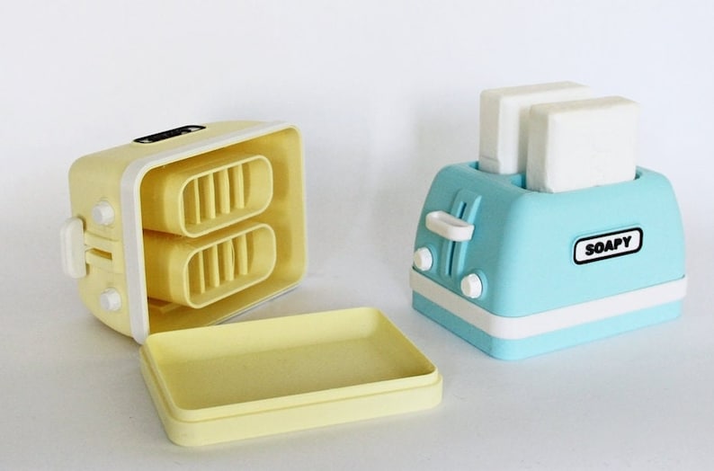 K&ouml;nnte beinhalten: Zwei dekorative Seifenschalen in Form von Toaster. Einer ist gelb und offen, so dass die Seife im Inneren sichtbar ist. Der andere ist blau und geschlossen mit zwei wei&szlig;en Seifenst&uuml;cken im Inneren. Der blaue Toaster hat den Schriftzug "SOAPY" auf der Vorderseite.