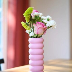 May include: A pink, modern vase with a stacked, rounded design holds a bouquet of fresh flowers. The arrangement includes pink roses, white carnations, and green chrysanthemums. The vase is on a wooden table, with a red curtain in the background.