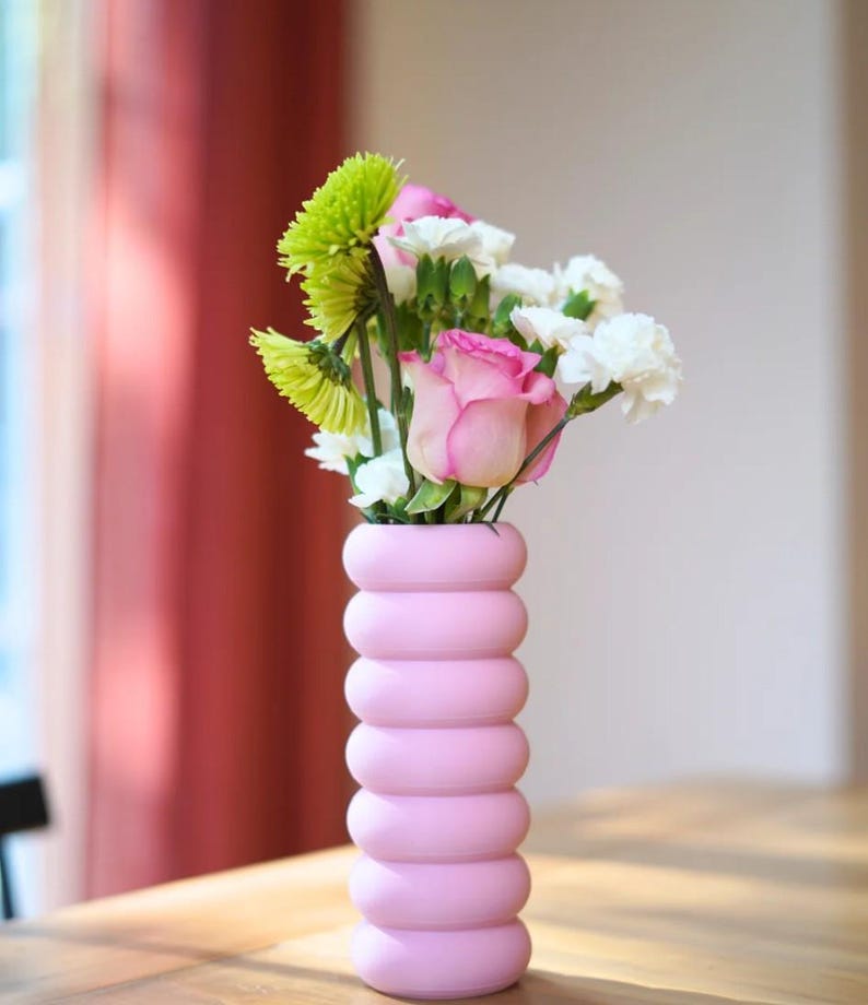 May include: A pink, modern vase with a stacked, rounded design holds a bouquet of fresh flowers. The arrangement includes pink roses, white carnations, and green chrysanthemums. The vase is on a wooden table, with a red curtain in the background.