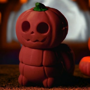 May include: A red, stacked pumpkin figurine with a carved jack-o'-lantern face. The figurine is sitting on a brown surface with other pumpkins in the background.