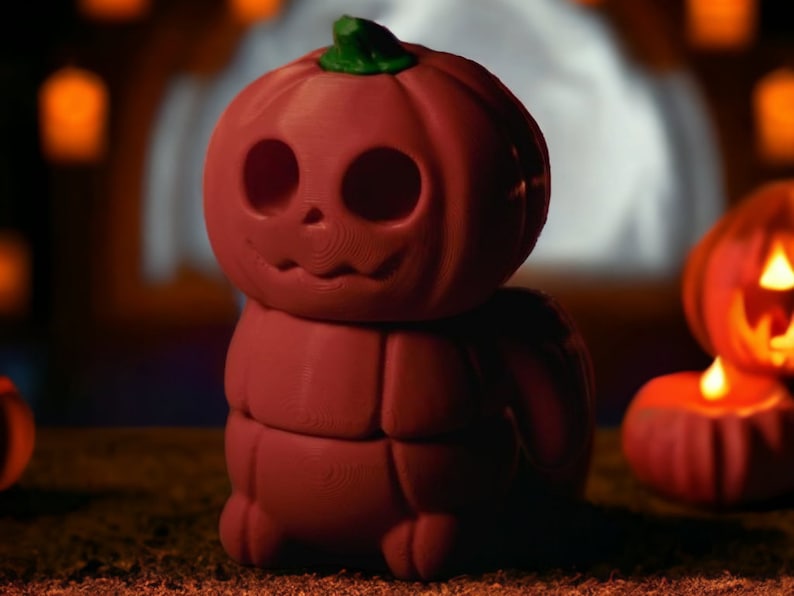 May include: A red, stacked pumpkin figurine with a carved jack-o'-lantern face. The figurine is sitting on a brown surface with other pumpkins in the background.