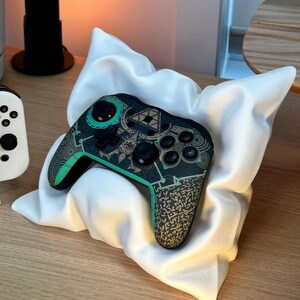 May include: A black and green video game controller with a decorative design featuring a green triangle and gold symbols. The controller is resting on a white pillow.